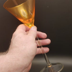 May include: A clear glass martini glass with an orange colored bowl. The glass is being held by a hand.