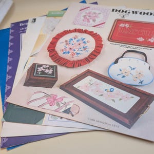 May include: A collection of cross-stitch patterns featuring floral designs. The patterns include a wooden box, a tray, a purse, and a welcome sign. The color palette includes pink, red, blue, and white. The designs are from Dogwoods.