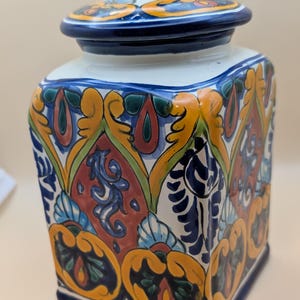 May include: A square ceramic jar with a lid, featuring a vibrant hand-painted design. The jar is adorned with intricate patterns in shades of blue, yellow, red, and green. The lid is dark blue with a small knob. A decorative kitchen storage item.