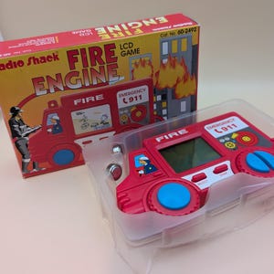 May include: A vintage Radio Shack Fire Engine LCD game in its original box. The red game console is shaped like a fire engine with blue wheels and a small screen. The box features a firefighter illustration and the words "Fire Engine".