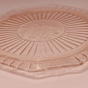 May include: A pink glass cake plate with a scalloped edge and a floral design in the center. The plate has a sunburst pattern radiating from the center.