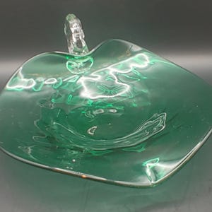 May include: A green glass dish with a clear glass handle. The dish is shaped like a leaf and has a wavy, textured surface.