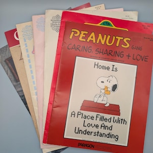 May include: A collection of vintage cross-stitch pattern books. The red cover displays Snoopy and the text "PEANUTS GANG CARING, SHARING + LOVE." Other covers are in shades of pink and beige, with floral and geometric patterns. The text "Home Is A Place Filled With Love And Understanding" is visible.