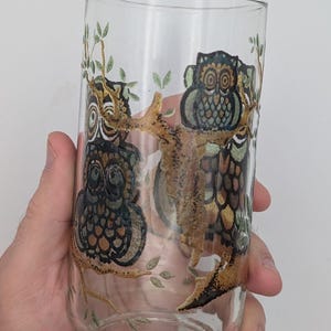 May include: Clear glass tumbler featuring a hand-painted design of three owls perched on a tree branch. The owls are depicted in shades of brown, green, and blue, with intricate details. The glass is held in a hand.