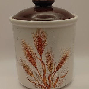 Puede incluir: Un tarro de cerámica blanco con tapa marrón y un diseño de espigas de trigo en tonos naranja y amarillo.