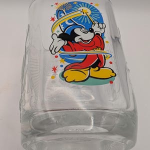 Puede incluir: Una taza de vidrio transparente con una imagen colorida de Mickey Mouse como mago con una varita y una estrella. El fondo es azul con estrellas amarillas y fuegos artificiales.