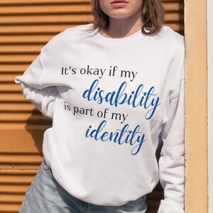 Könnte beinhalten: Weißes Sweatshirt mit dem Text "It's okay if my disability is part of my identity" in schwarzer und blauer Schrift. Die Person trägt eine Sonnenbrille und Jeansshorts. Der Hintergrund ist eine strukturierte orangefarbene Wand.