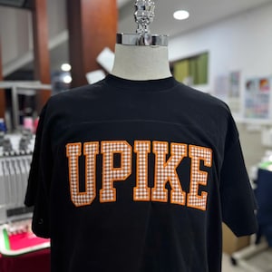 May include: Black t-shirt with the word "UPIKE" in orange and white gingham letters. The shirt is displayed on a mannequin. The t-shirt has a crew neck and short sleeves.