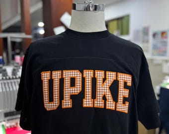 May include: Black t-shirt with the word "UPIKE" in orange and white gingham letters. The shirt is displayed on a mannequin. The t-shirt has a crew neck and short sleeves.