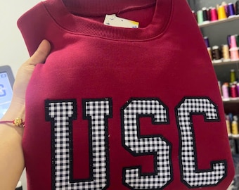 May include: A maroon sweatshirt with the letters "USC" in a black and white checkered pattern. The sweatshirt is held up by a hand, and the background is blurred.
