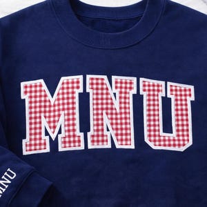 May include: Navy blue sweatshirt with the letters "MNU" in a red and white gingham pattern across the chest. The left sleeve has "MNU" embroidered in white. A casual, comfortable top.