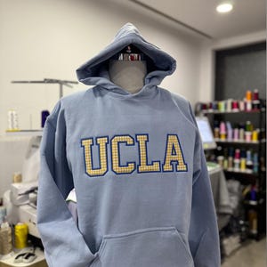 Puede incluir: Sudadera azul claro de la UCLA con capucha y bolsillo delantero. Las letras "UCLA" est&aacute;n bordadas en oro y cuadros azules. La sudadera se exhibe en un maniqu&iacute;.