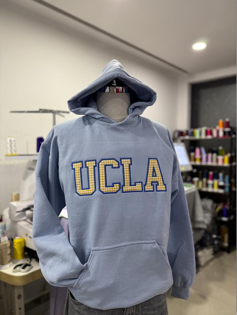 Puede incluir: Sudadera azul claro de la UCLA con capucha y bolsillo delantero. Las letras "UCLA" est&aacute;n bordadas en oro y cuadros azules. La sudadera se exhibe en un maniqu&iacute;.