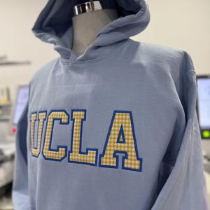 Puede incluir: Sudadera con capucha azul claro con las letras "UCLA" bordadas en la parte delantera. Las letras son amarillas con un estampado de cuadros y contornos azules. La sudadera tiene capucha y se muestra en un maniqu&iacute;.