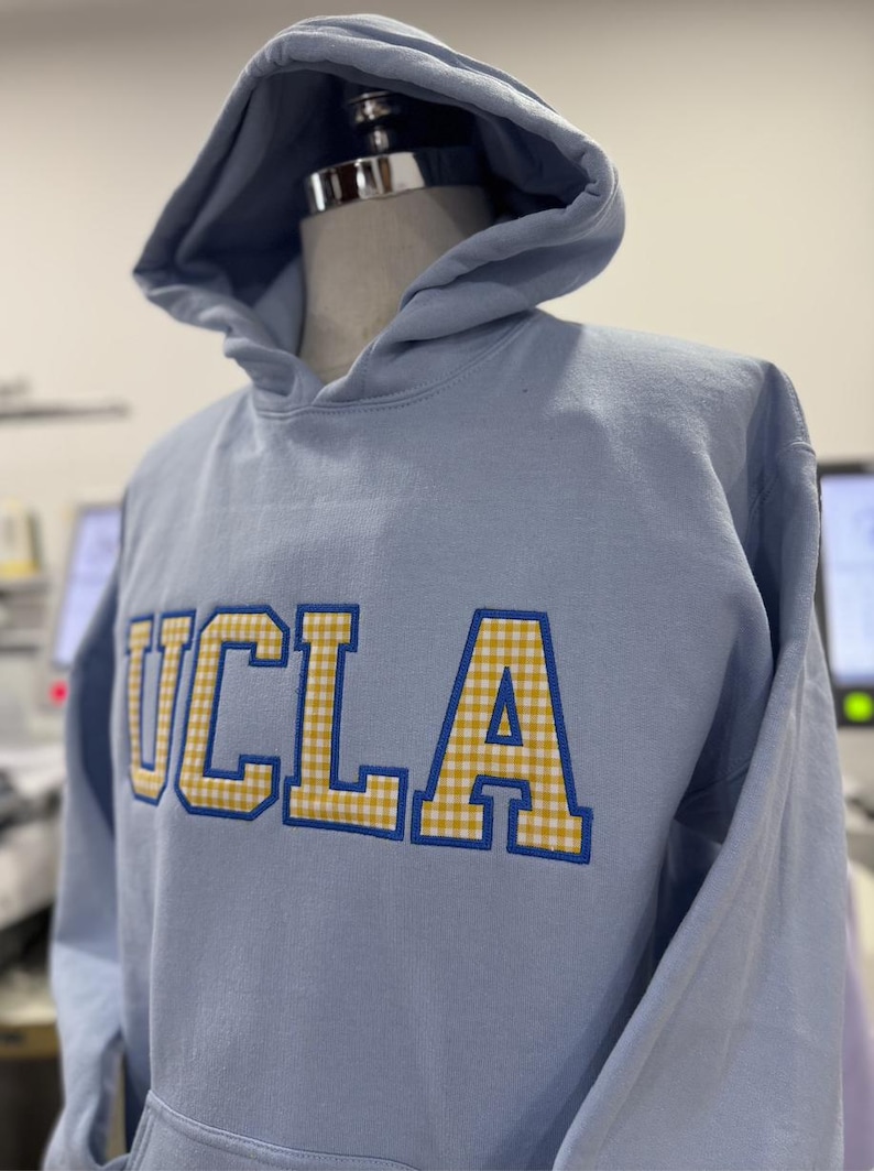 Puede incluir: Sudadera con capucha azul claro con las letras "UCLA" bordadas en la parte delantera. Las letras son amarillas con un estampado de cuadros y contornos azules. La sudadera tiene capucha y se muestra en un maniqu&iacute;.