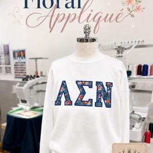 Puede incluir: Sudadera blanca de cuello redondo con letras florales aplicadas. Las letras, en azul con un estampado floral, deletrean una combinaci&oacute;n de letras griegas. Las palabras "Floral Appliqu&eacute;" est&aacute;n escritas encima de la sudadera en cursiva.