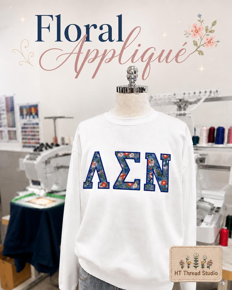 Puede incluir: Sudadera blanca de cuello redondo con letras florales aplicadas. Las letras, en azul con un estampado floral, deletrean una combinaci&oacute;n de letras griegas. Las palabras "Floral Appliqu&eacute;" est&aacute;n escritas encima de la sudadera en cursiva.
