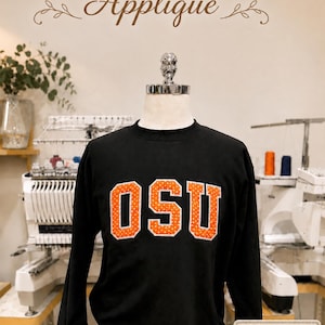 Puede incluir: Sudadera negra de manga larga con las letras "OSU" en naranja con lunares blancos. El texto "Polka Dot Appliqu&eacute;" est&aacute; encima de la sudadera. La sudadera se muestra en un maniqu&iacute;.