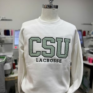 Puede incluir: Sudadera blanca con el texto "CSU LACROSSE" en verde y negro. Las letras "CSU" tienen un patr&oacute;n de cuadros verdes y blancos. La sudadera se muestra en un maniqu&iacute;.