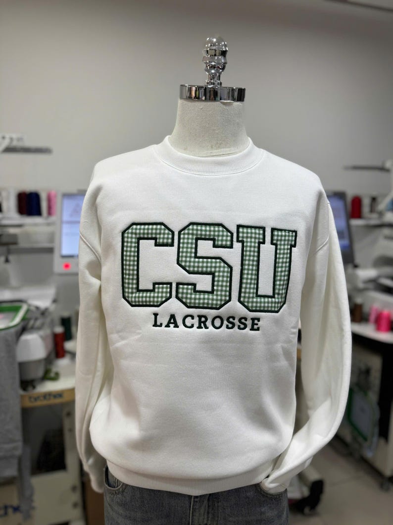 Puede incluir: Sudadera blanca con el texto "CSU LACROSSE" en verde y negro. Las letras "CSU" tienen un patr&oacute;n de cuadros verdes y blancos. La sudadera se muestra en un maniqu&iacute;.