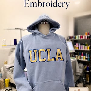 Puede incluir: Una sudadera con capucha azul claro con bolsillo delantero y capucha, exhibida en un maniqu&iacute;. La palabra "UCLA" est&aacute; bordada en el pecho en cuadros amarillos y dorados. El texto "Gingham Appliqu&eacute; Embroidery" est&aacute; encima de la sudadera.