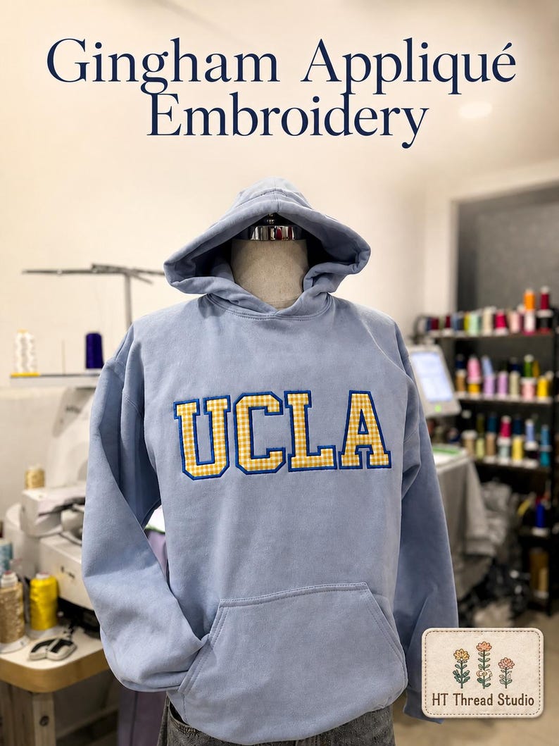 Puede incluir: Una sudadera con capucha azul claro con bolsillo delantero y capucha, exhibida en un maniqu&iacute;. La palabra "UCLA" est&aacute; bordada en el pecho en cuadros amarillos y dorados. El texto "Gingham Appliqu&eacute; Embroidery" est&aacute; encima de la sudadera.