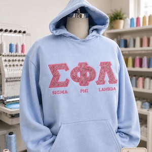 Puede incluir: Una sudadera con capucha azul claro con un bolsillo delantero y una capucha. La parte delantera de la sudadera con capucha presenta las letras griegas Sigma, Phi y Lambda en una aplicaci&oacute;n rosa brillante. El texto "Sigma Phi Lambda" est&aacute; debajo. El texto "Glitter Appliqu&eacute; Embroidery Style" est&aacute; en la parte superior.