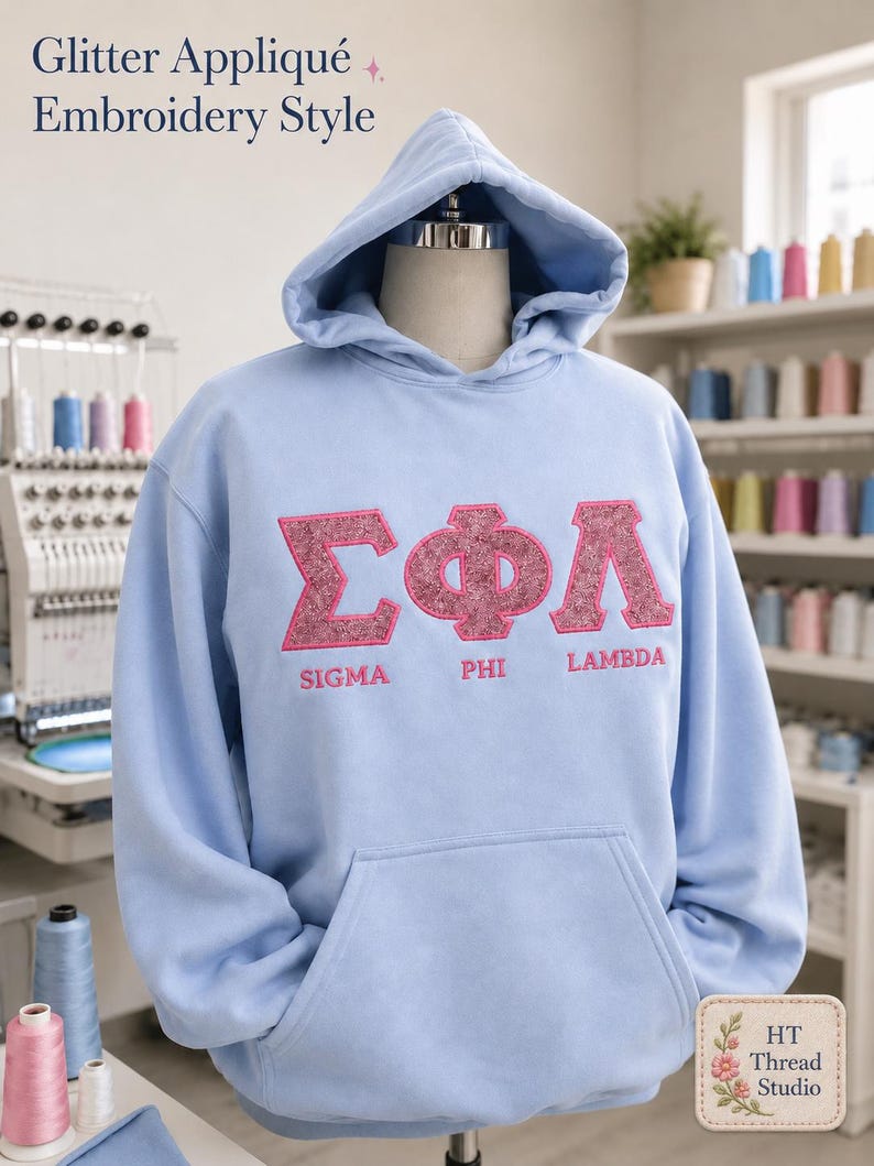 Puede incluir: Una sudadera con capucha azul claro con un bolsillo delantero y una capucha. La parte delantera de la sudadera con capucha presenta las letras griegas Sigma, Phi y Lambda en una aplicaci&oacute;n rosa brillante. El texto "Sigma Phi Lambda" est&aacute; debajo. El texto "Glitter Appliqu&eacute; Embroidery Style" est&aacute; en la parte superior.