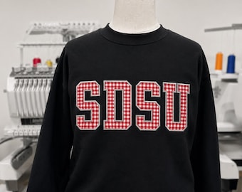 May include: Black crew neck sweatshirt with the letters "SDSU" in a red and white gingham pattern. The sweatshirt is displayed on a mannequin. The background is a light gray.