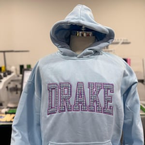 Puede incluir: Una sudadera con capucha azul claro con el nombre "DRAKE" bordado en el pecho en un patr&oacute;n de cuadros morado y negro. La sudadera con capucha tiene una capucha y un bolsillo delantero. La prenda se exhibe en un maniqu&iacute;.