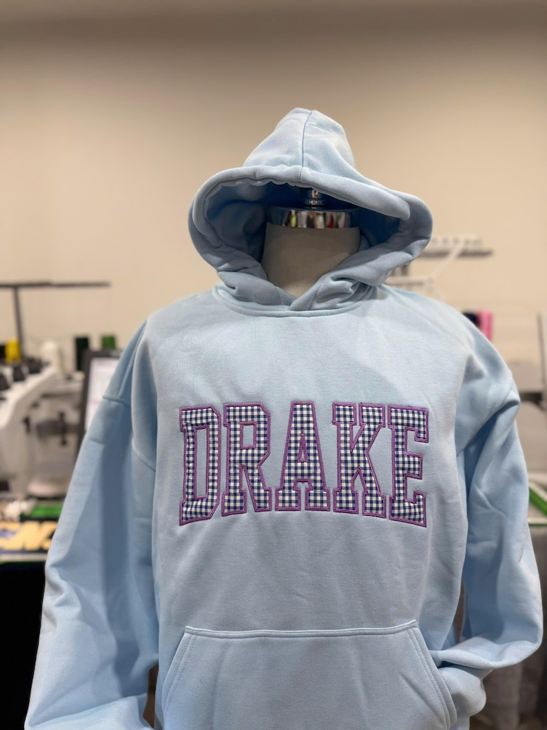 Puede incluir: Una sudadera con capucha azul claro con el nombre "DRAKE" bordado en el pecho en un patr&oacute;n de cuadros morado y negro. La sudadera con capucha tiene una capucha y un bolsillo delantero. La prenda se exhibe en un maniqu&iacute;.