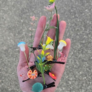 May include: A collection of decorative hair clips featuring various designs, including flowers, mushrooms, and small animal figures. The clips are in various colors, such as pink, yellow, and blue, and are arranged together.