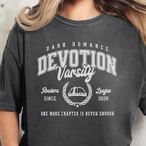 May include: A charcoal gray short-sleeve t-shirt featuring white screen-printed text that reads Dark Romance Devotion Varsity, Readers League Since 2026, and One More Chapter Is Never Enough, with a central graphic of an open book and laurel leaves.