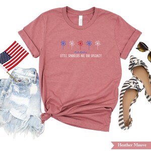 May include: A heather mauve t-shirt with a graphic of red, white, and blue fireworks and the text "Little sparklers are our specialty".