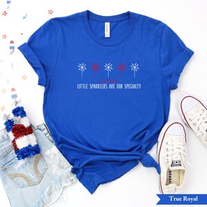 May include: A royal blue t-shirt with a red, white, and blue firework design and the text "Little Sparklers Are Our Specialty".