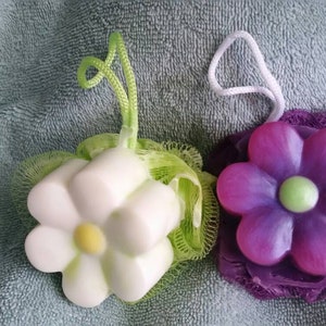 May include: Two flower-shaped bath soaps, one white with a yellow center and one purple with a green center, are wrapped in mesh bags with string handles.