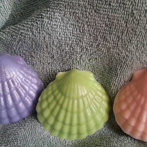 May include: Three decorative seashell-shaped soaps in purple, green, and pink. The soaps have a shimmery finish and are arranged in a row on a gray textured surface.