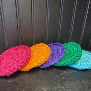 May include: A row of six colorful, oval-shaped crochet items in shades of pink, orange, purple, green, and blue. The items are arranged in a line, showcasing their textured, handmade appearance. The background is a dark wooden surface.
