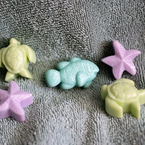 May include: Five handmade soap bars shaped like sea creatures. There are two green turtle soaps, a blue fish soap, and two purple starfish soaps.