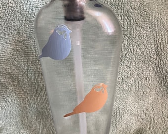 May include: A clear plastic pump bottle with a black pump top. The bottle has three bird decals on it: one blue bird, one orange bird, and one blue bird.