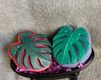 May include: Two green and purple leaf-shaped soaps in a black bathtub-shaped soap dish. The soaps are surrounded by purple shredded paper.