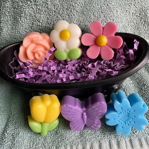 May include: A black bowl filled with purple paper shreds, holding six colorful, flower-shaped soaps. The soaps include a peach rose, a white daisy with green leaves, a pink flower, a yellow tulip, a purple butterfly, and a blue flower.
