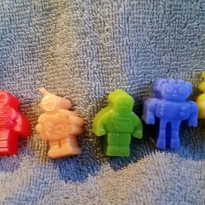 May include: Six small robot figurines made of wax. The robots are in a rainbow color scheme, starting with red, then orange, green, blue, yellow, and ending with purple.