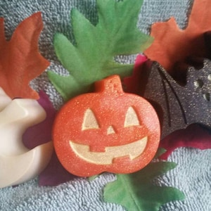 May include: A Halloween-themed arrangement featuring a smiling ghost, a carved pumpkin, and a bat. The ghost is off-white, the pumpkin is orange, and the bat is dark gray. Colorful leaves surround the figures, creating a festive display.