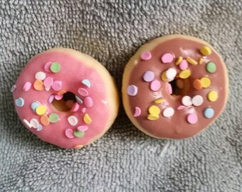May include: Two donuts with colorful sprinkles. One donut is pink with white sprinkles and the other is brown with white, yellow, and blue sprinkles.