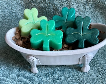 May include: Four green shamrock-shaped bath bombs in a white bathtub-shaped dish. The bath bombs are different shades of green.