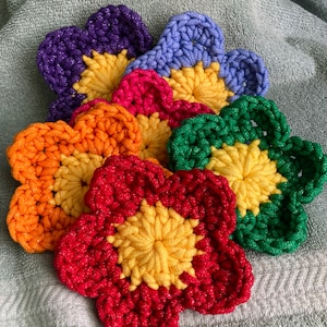 May include: A collection of colorful, crocheted flower-shaped items. The flowers feature petals in shades of red, orange, green, pink, purple, and blue, each with a yellow center. The items are handmade and appear to be coasters or decorative pieces.