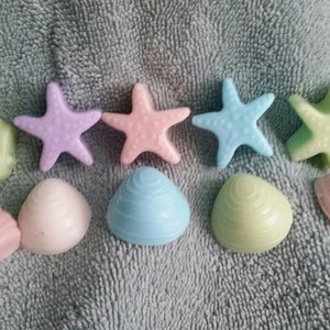 May include: Assortment of pastel-colored seashell and starfish shaped soaps. The soaps are arranged on a light gray textured surface.