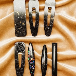 May include: A collection of black hair clips and barrettes with various designs. Some feature gold flecks, others have a gradient effect, and some are adorned with colorful glitter or a rose design. The clips are displayed on a gold satin surface.