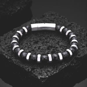 Puede incluir: Una pulsera de cuentas que alterna cuentas negras facetadas y espaciadores de metal plateado pulido. La pulsera incluye un cierre magnético rectangular de metal plateado grabado con el texto NAYA-LINN en una fuente sans-serif clara.